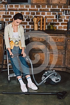 sad young repairwoman looking at broken vacuum cleaner while sitting on stepladder