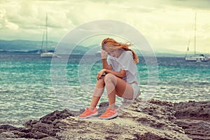 Sad young serious woman sitting on the rock looking down with sea ocean sky background