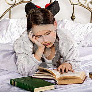 Sad young student girl reads a book while lying on a bed doing homework