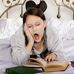 Sad young student girl reads a book while lying on a bed doing homework