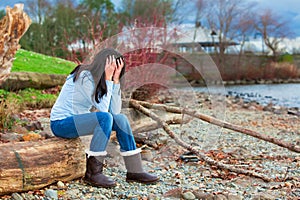 Sad young teen girl sitting on log along rocky beach by lake