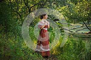 Sad young woman in a 19th century dress by the river with a book in her hands. Summer landscape. The photo