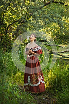 Sad young woman in a 19th century dress by the river with a book in her hands. Summer landscape. The photo