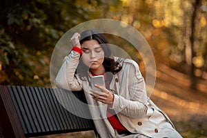Sad young woman in autumn outfit sitting on bench at park with smartphone and feeling depressed