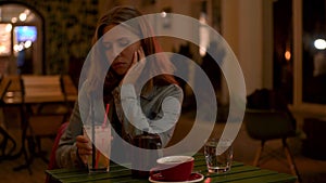 Sad young woman with brown hair sitting along at the table in a cafe and drinking a cocktail. Media. Lonely girl waiting