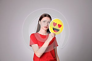 Sad young woman covering face with heart eyes emoji on grey background