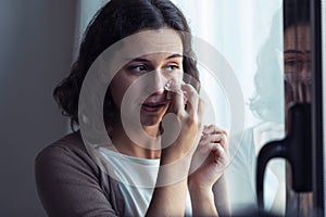 Sad young woman crying while looking through the window at home