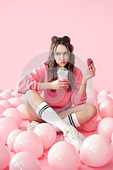 Sad young woman eating milkshake and donut sitting on floor with many pink air balloons