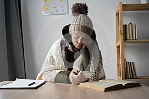 Sad young woman sit at the table at freezing cooled flat in warm cap and blanket shiver tremble with cold, using cell