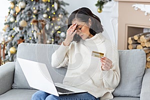 Sad young woman sitting at home on the sofa for the holidays near the Christmas tree, holding a credit card and holding