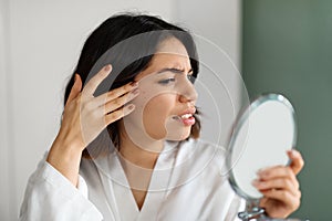 Sad young woman suffering from acne, checking skin in mirror