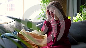 A sad young woman is suffering from a severe headache. Curly woman sits on the couch and grimaces from pain in the