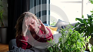 A sad young woman is suffering from a severe headache. Curly woman sits on the couch and grimaces from pain in the