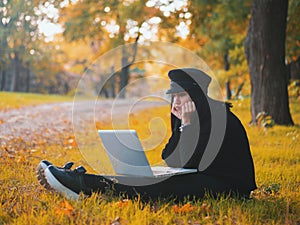 Sad young woman using laptop outdoor sitting on yellow grass. Girl using digital tablet pc in the autumn park. Trendy