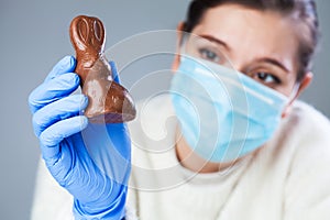 Sad young woman wearing protective face mask and medical gloves holding a chocolate Easter bunny