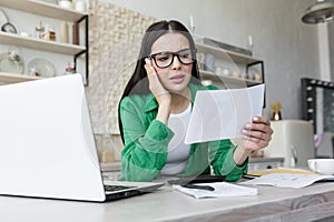 Sad young woman working from home with laptop and documents. Holds in hands and anxiously looks at bills, letter