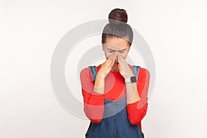 Sadness and depression. Portrait of upset girl with hair bun in denim overalls bowing her head and crying