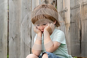 Sadness. Sad boy. A lonely child sits near a wooden fence. Frustration. Sad emotions. Experiences. Child psychology