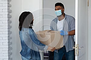 Safe Delivery Service. Black courier wearing mask giving cardboard box to woman