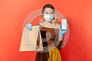 Safe online shopping on quarantine. Woman in protective mask and gloves holding package with food and mobile phone