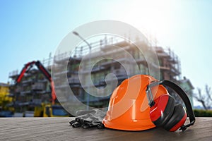 Safety equipment. Hard hat, gloves and protective headphones on wooden surface near unfinished building outdoors, space for text