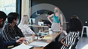 Safety measures during pandemic at work. Young Caucasian business woman leading office meeting, employees wearing masks.