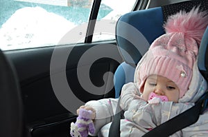 Safety seat for baby in car. Little cute girl in a pink hat and overalls sits winter in a child fastened with straps.