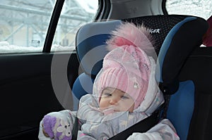 Safety seat for baby in car. Little cute girl in a pink hat and overalls sits winter in a child fastened with straps.
