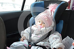 Safety seat for baby in car. Little cute girl in a pink hat and overalls sits winter in a child fastened with straps.