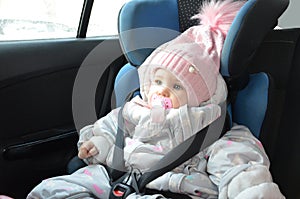 Safety seat for baby in car. Little cute girl in a pink hat and overalls sits winter in a child fastened with straps.