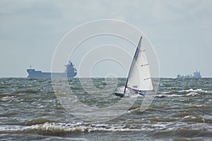 A sailboat makes pace along the beach. Large freighters on the horizon.