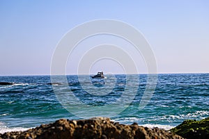 A sailboat in the middle of vast blue ocean water with red flowers and lush green plants on the hillside with blue sky