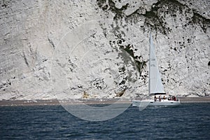 A sailboat motoring along on the sea under white cliffs close to the shore