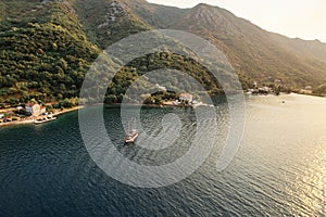 Sailboat sails across the bay against the backdrop of a green, wooded mountain coast. Drone