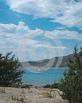 Sailboat seen from between green bushes on in Elafonisos, Greece on a beautiful day with blue sky and clouds. Simos beach.
