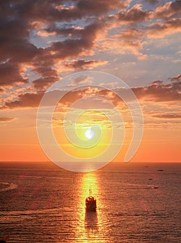 A sailing boat towards the sun, aligned with the sun reflection on water during a slightly cloudy sunset