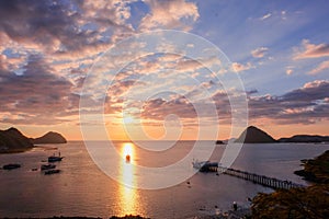 A sailing boat towards the sun, aligned with the sun reflection on water during a slightly cloudy sunset with a marina in Labuan