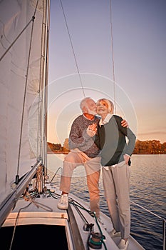 Sailing at sunset. Full length of happy senior couple holding hands and hugging while standing on the side of yacht deck