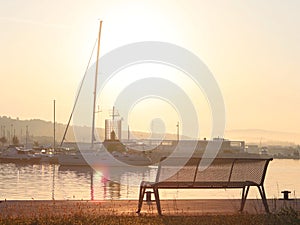 The sailing yacht enters under the motor in the marina in the rays of the morning sun. Dawn in the port. Empty bench on the pier a