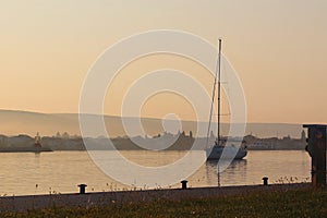 The sailing yacht enters under the motor in the marina in the rays of the morning sun. Dawn in the port. Empty pier with