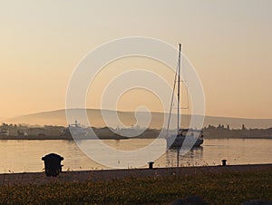 The sailing yacht enters under the motor in the marina in the rays of the morning sun. Dawn in the port. Empty pier with electrica