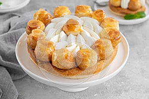 Saint Honore cake with profitrols, caramel, custard and whipped cream on a white plate on a gray concrete background.
