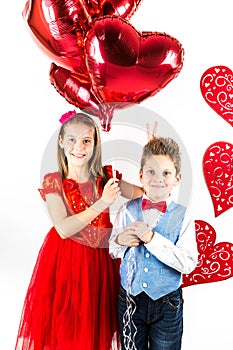 Saint Valentine`s day. Pretty girl with red dress and gentleman boy with blue vest, red butterfly tie, red roses bucket and heart