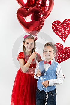 Saint Valentine`s day. Pretty girl with red dress and gentleman boy with blue vest, red butterfly tie, red roses bucket and heart