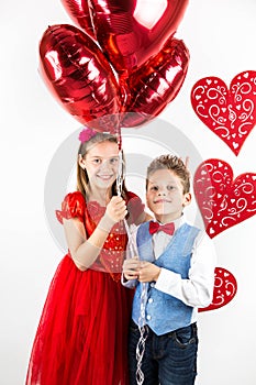 Saint Valentine`s day. Pretty girl with red dress and gentleman boy with blue vest, red butterfly tie, red roses bucket and heart