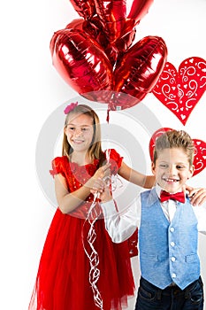 Saint Valentine`s day. Pretty girl with red dress and gentleman boy with blue vest, red butterfly tie, red roses bucket and heart