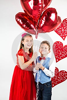 Saint Valentine`s day. Pretty girl with red dress and gentleman boy with blue vest, red butterfly tie, red roses bucket and heart