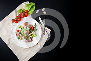 Salad with beef, cheese, herbs and quail egg on a white plate next to a fork and knife. on the black table. decorated with a