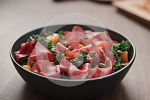 Salad with kale, bresaola and cherry tomatoes decorated with capers in black bowl on walnut wood table