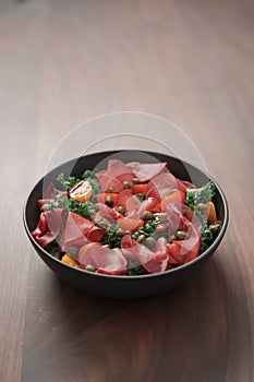 Salad with kale, bresaola and cherry tomatoes decorated with capers in black bowl on walnut wood table